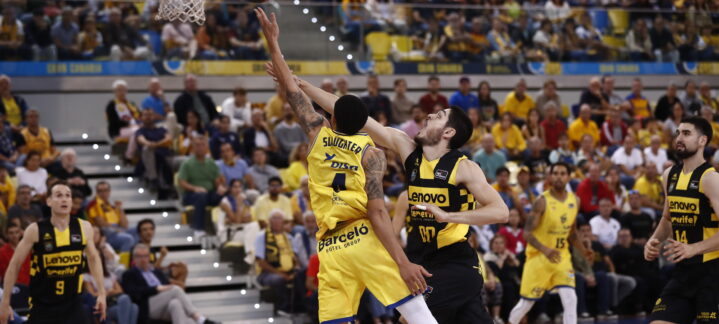 Jugadores compiten en un partido de baloncesto, con el público animando de fondo.