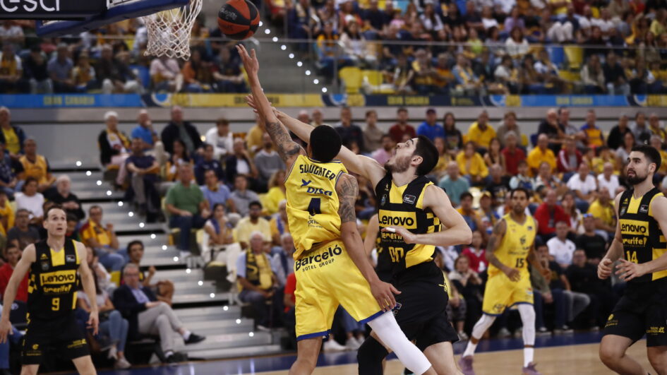 Jugadores compiten en un partido de baloncesto, con el público animando de fondo.