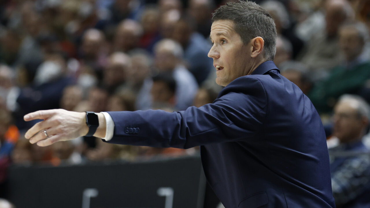 Entrenador dando instrucciones durante un partido de baloncesto, público al fondo.