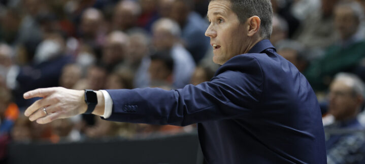 Entrenador dando instrucciones durante un partido de baloncesto, público al fondo.