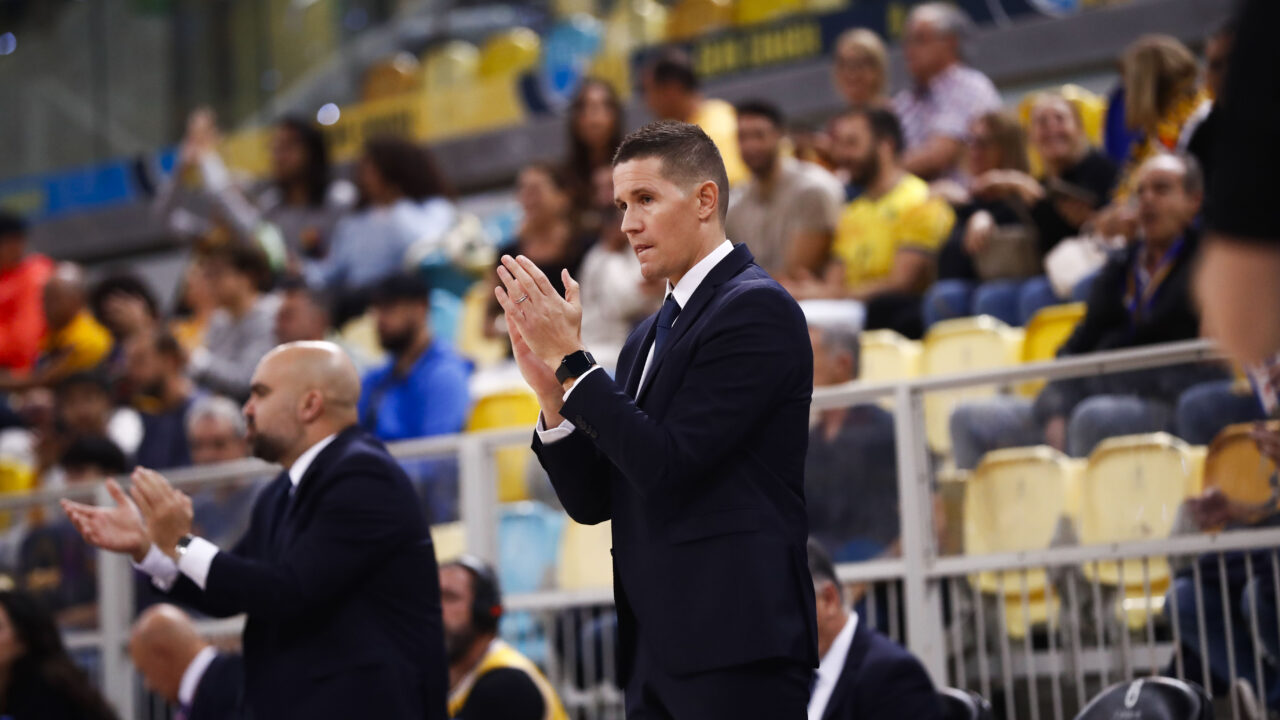 Entrenador aplaudiendo en un partido, con afición al fondo en un ambiente animado.