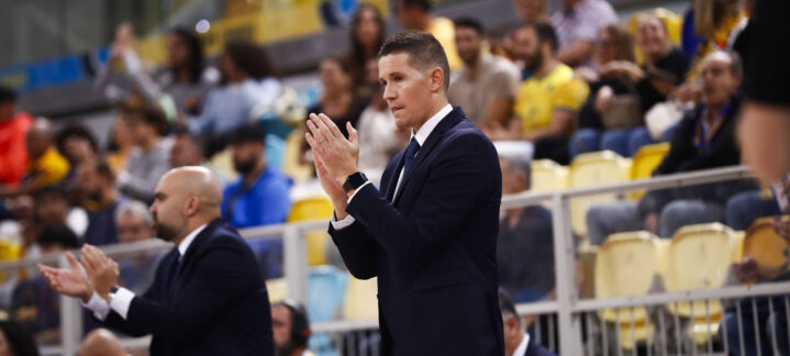 Entrenador aplaudiendo en un partido, con afición al fondo en un ambiente animado.