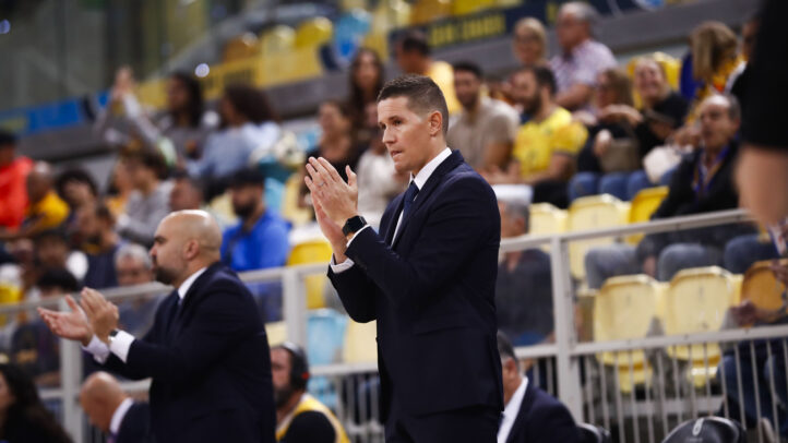 Entrenador aplaudiendo en un partido, con afición al fondo en un ambiente animado.