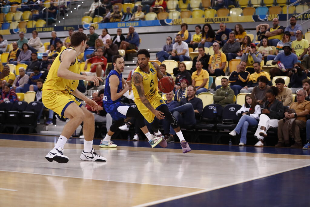 Jugadores de baloncesto en acción durante un partido en el estadio, con público animando.