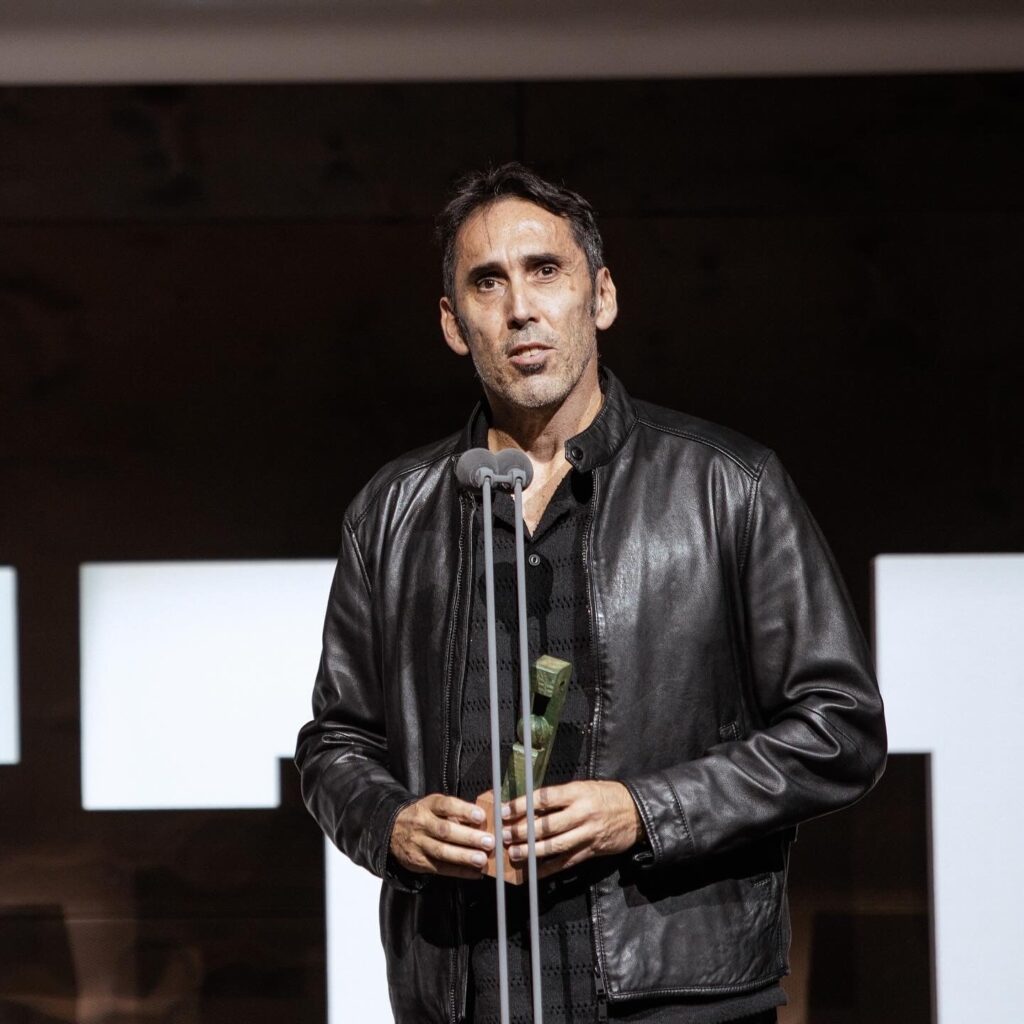 Hombre en traje de cuero pronuncia un discurso sosteniendo un premio en un evento.
