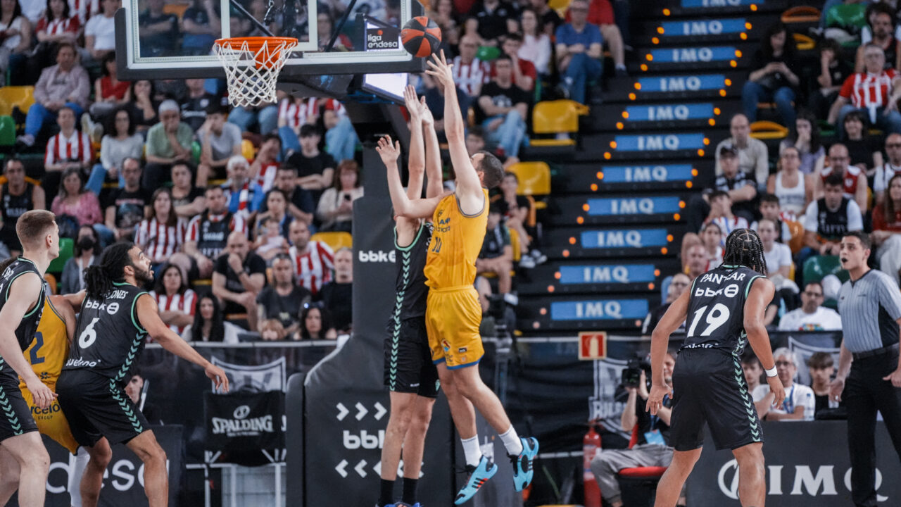 Jugadores compiten por un rebote en un partido de baloncesto en un ambiente animado.