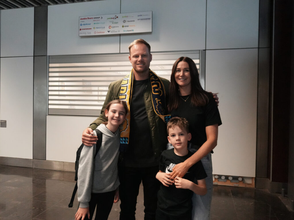 en la isla para su homenaje: ""Es genial estar en casa""","Brad Newley posando con su familia en el aeropuerto, envuelto en una bufanda del equipo.