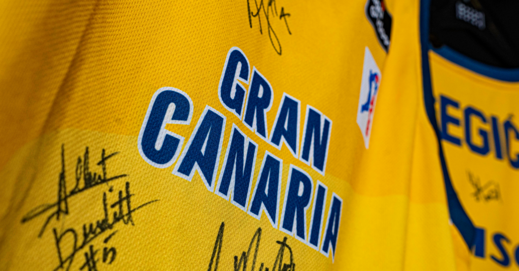  a subasta con fines benéficos",Camiseta amarilla del Gran Canaria con múltiples firmas de jugadores.