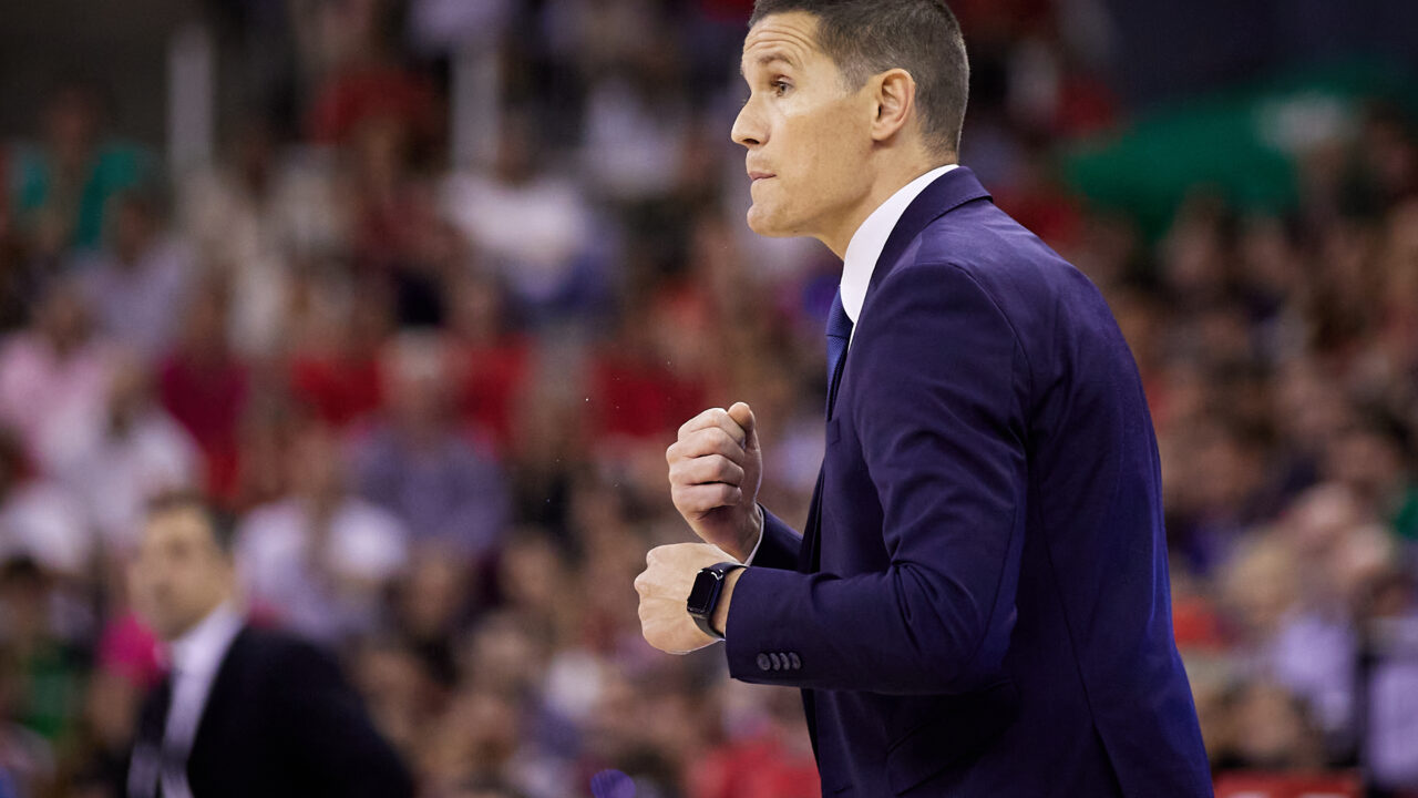 Entrenador gesticulando en un partido de baloncesto, con afición al fondo.
