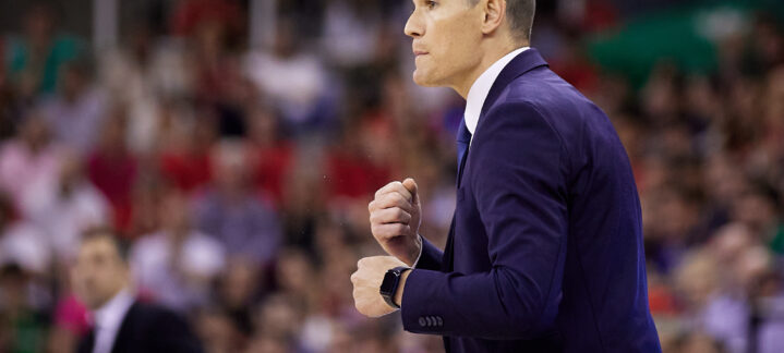 Entrenador gesticulando en un partido de baloncesto, con afición al fondo.