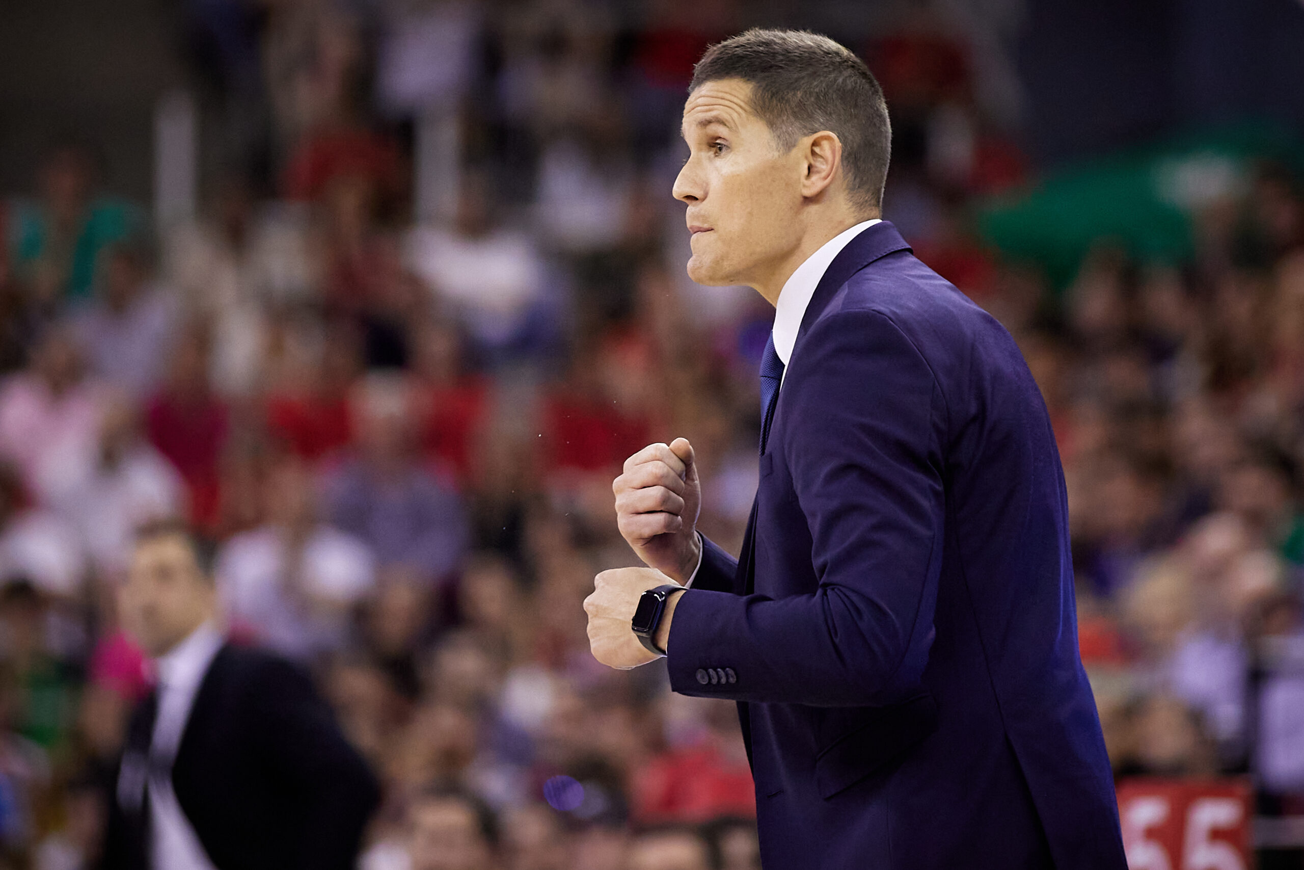 Entrenador gesticulando en un partido de baloncesto, con afición al fondo.