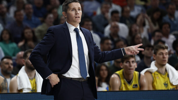 Entrenador gesticulando durante un partido, con jugadores en la banca al fondo.
