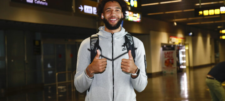 Jugador sonriente en aeropuerto, con pulgar hacia arriba y mochila, listo para unirse al equipo.