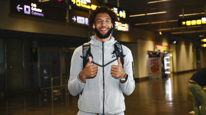 Jugador sonriente en aeropuerto, con pulgar hacia arriba y mochila, listo para unirse al equipo.