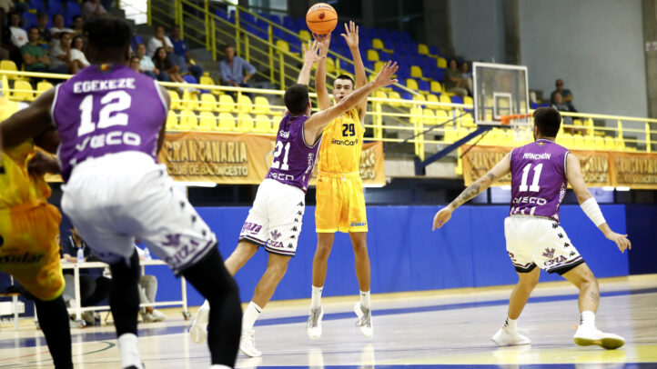 Jugador lanzando un balón de baloncesto mientras es defendido por dos contrincantes.