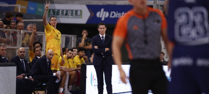 Entrenador observa el partido mientras un jugador hace un gesto de victoria desde el banquillo.