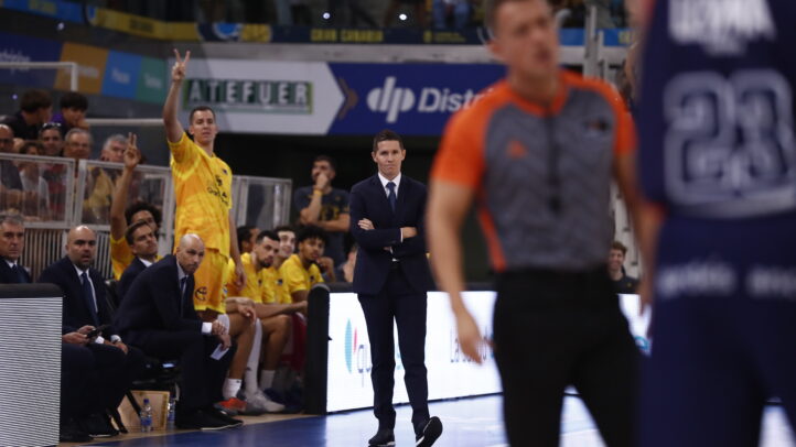 Entrenador observa el partido mientras un jugador hace un gesto de victoria desde el banquillo.
