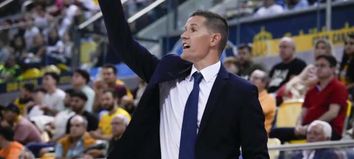 Entrenador dando instrucciones en un partido de baloncesto, con público al fondo.