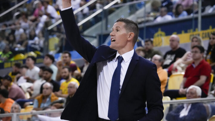 Entrenador dando instrucciones en un partido de baloncesto, con público al fondo.