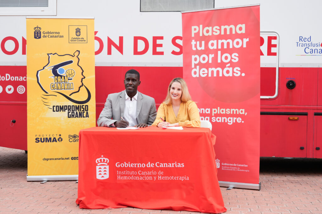 Dos personas firman un acuerdo en un evento de donación de sangre, junto a banners promocionales.