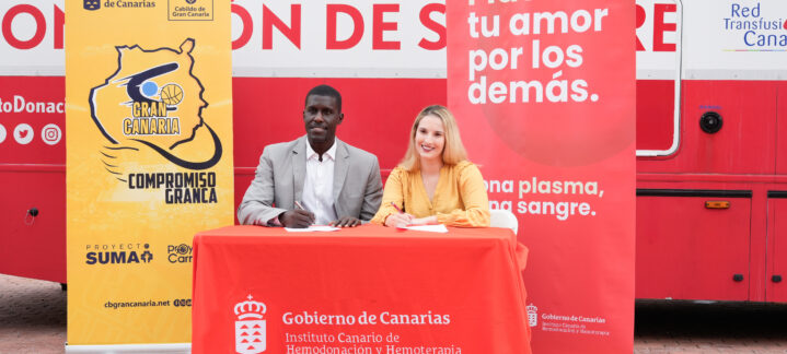 Dos personas firman un acuerdo en un evento de donación de sangre, junto a banners promocionales.