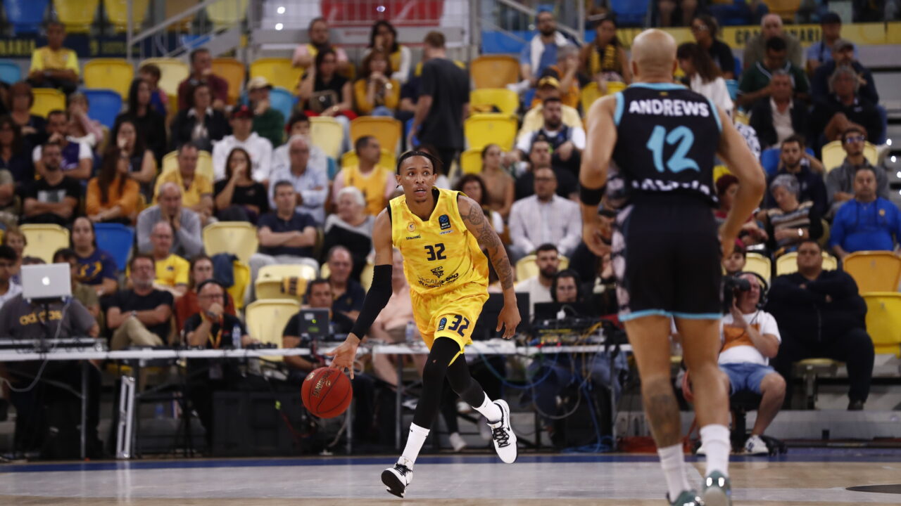 Jugador con balón en un partido de baloncesto, con afición al fondo.