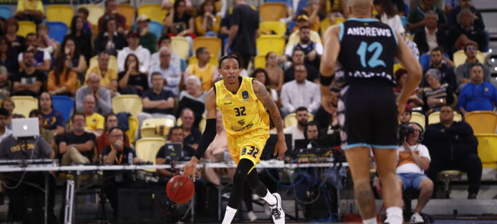 Jugador con balón en un partido de baloncesto, con afición al fondo.