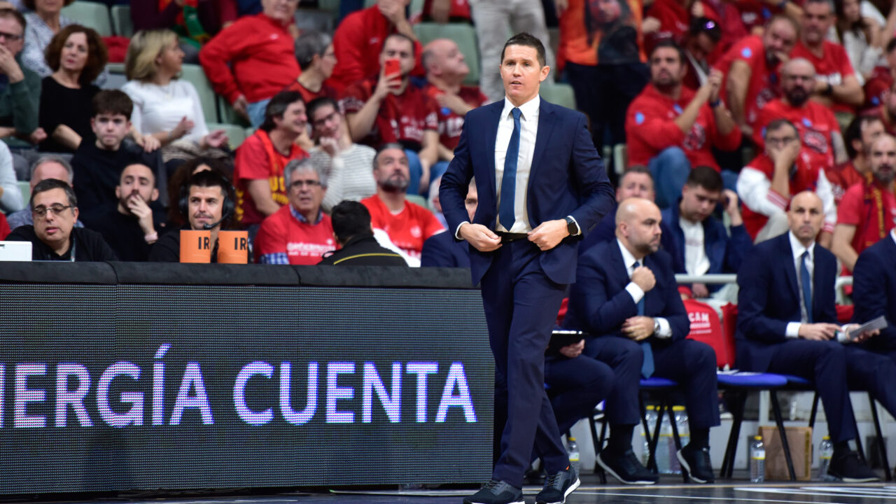 Entrenador Jaka Lakovic dirigiendo el partido con aficionados animando en el fondo.