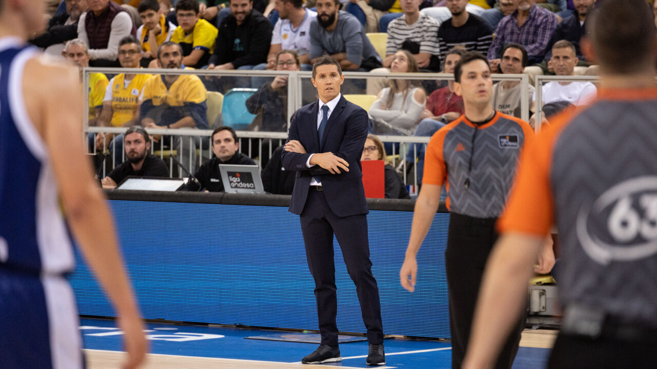 Entrenador en traje observa un partido de baloncesto con jugadores y árbitros en acción.