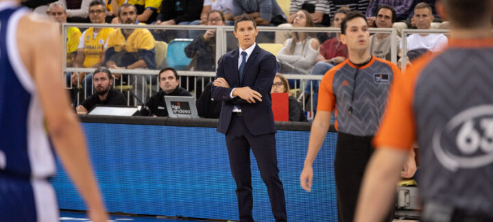 Entrenador en traje observa un partido de baloncesto con jugadores y árbitros en acción.