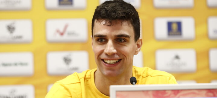 Jugador del CB Gran Canaria sonríe durante una rueda de prensa. Fondo amarillo con logotipos.
