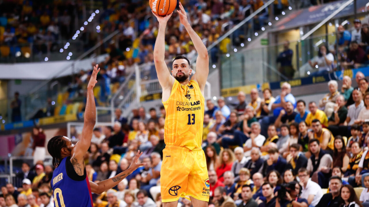 Jugador lanzando a canasta en un partido de baloncesto con público animando.