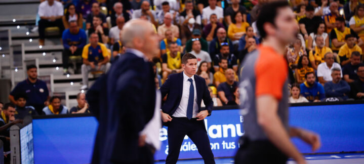 Entrenador junto a árbitros en un partido de baloncesto, con público animado al fondo.