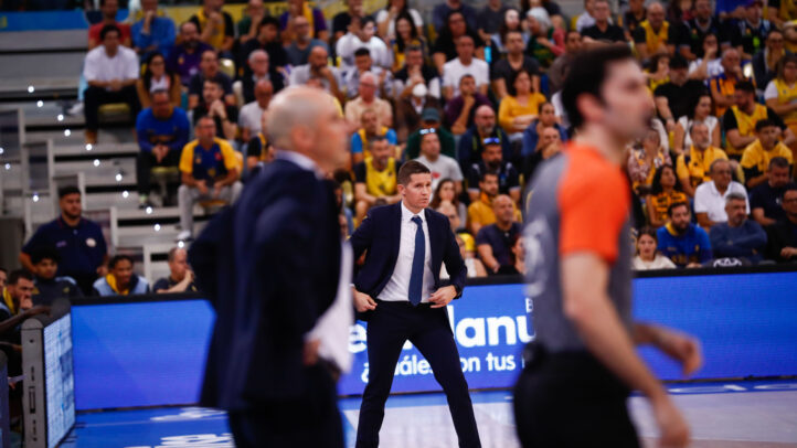 Entrenador junto a árbitros en un partido de baloncesto, con público animado al fondo.