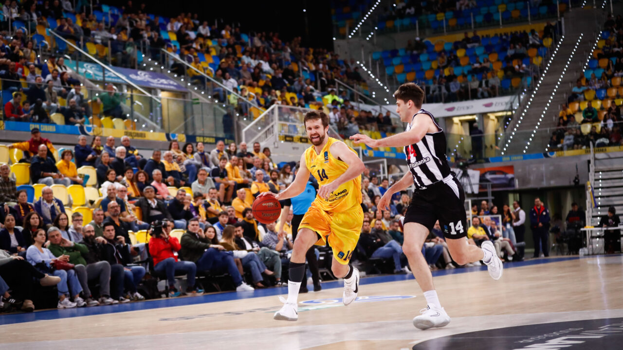 Jugadores de baloncesto compiten en un partido con una multitud animada de fondo.