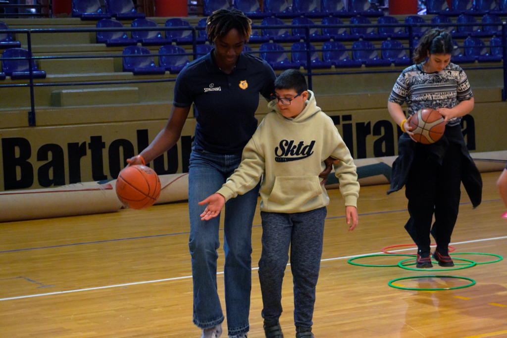 Jugadora de baloncesto guía a un niño en entrenamiento, rodeados de otros participantes.
