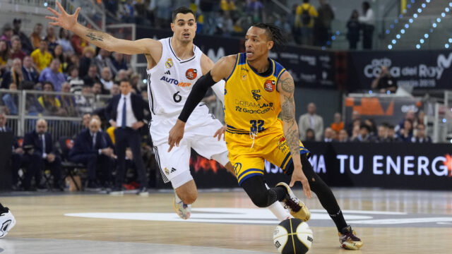 Dos jugadores compiten en un partido de baloncesto, uno con balón y el otro defendiendo.