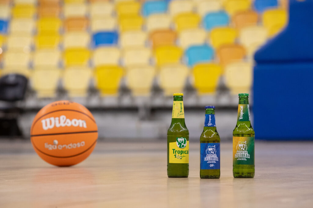 Botellas de cerveza Tropical frente a un balón de baloncesto en un ambiente deportivo.