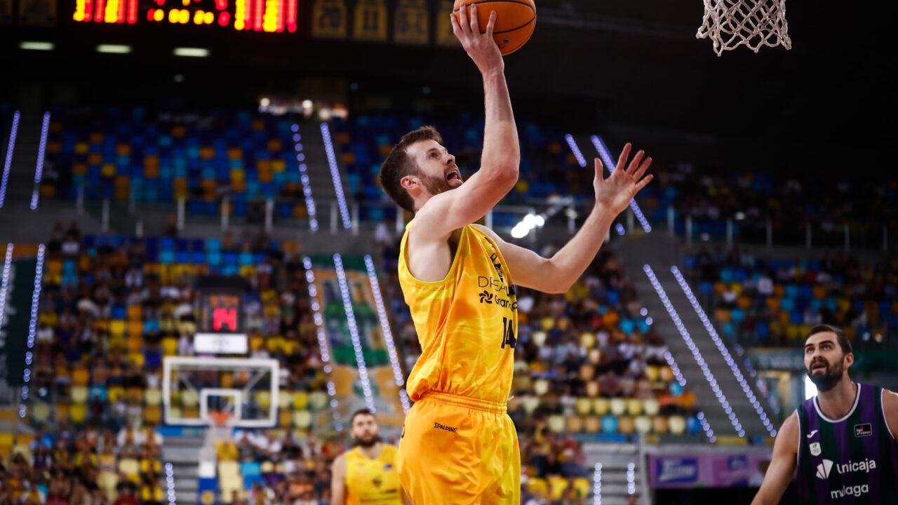 Jugador lanzando a canasta durante un partido de baloncesto. Público en las gradas.