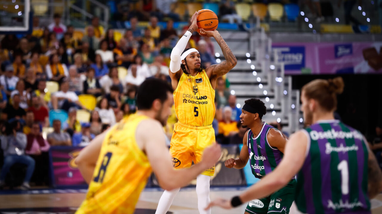 Jugador lanzando a canasta en un partido de baloncesto, con la afición al fondo.