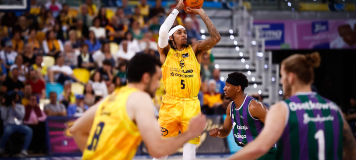 Jugador lanzando a canasta en un partido de baloncesto, con la afición al fondo.