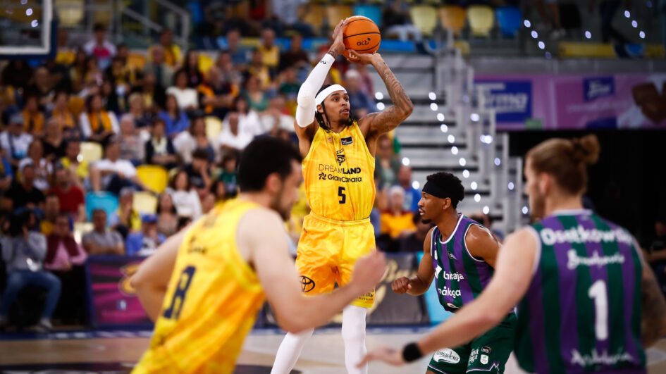 Jugador lanzando a canasta en un partido de baloncesto, con la afición al fondo.