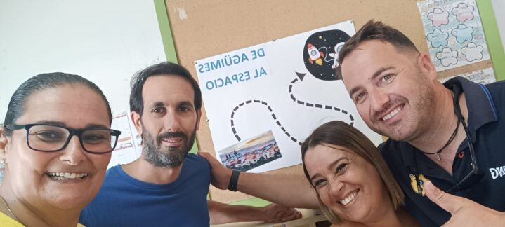 Cuatro personas sonrientes posan frente a un cartel sobre el espacio en un aula escolar.