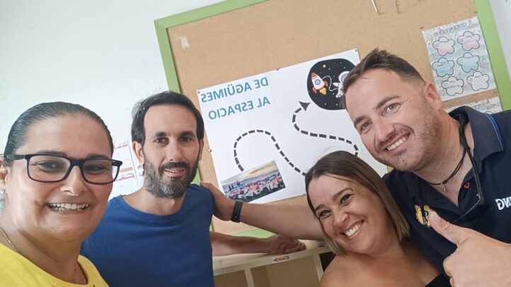 Cuatro personas sonrientes posan frente a un cartel sobre el espacio en un aula escolar.