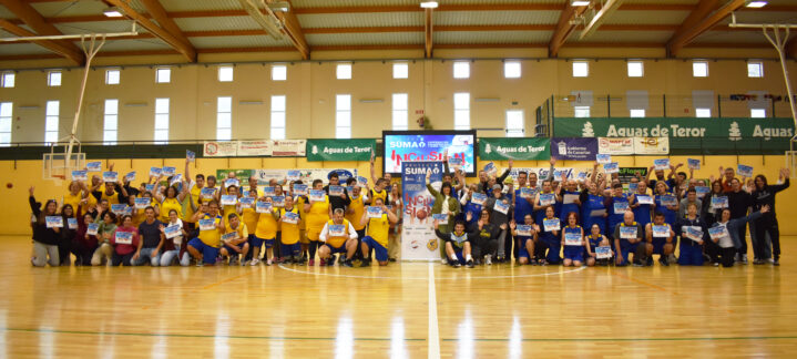 Grupo de personas sonrientes en un evento deportivo, sosteniendo carteles en un polideportivo.