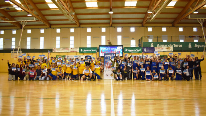 Grupo de personas sonrientes en un evento deportivo, sosteniendo carteles en un polideportivo.