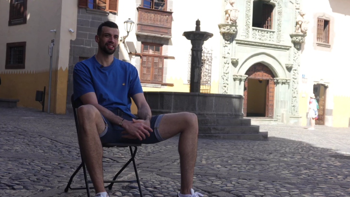 todos los días""","Pierre Pelos sentado en una silla en una plaza pintoresca, sonriendo.