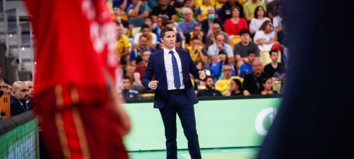 Entrenador en la cancha, gesticulando durante un partido, con aficionados al fondo.