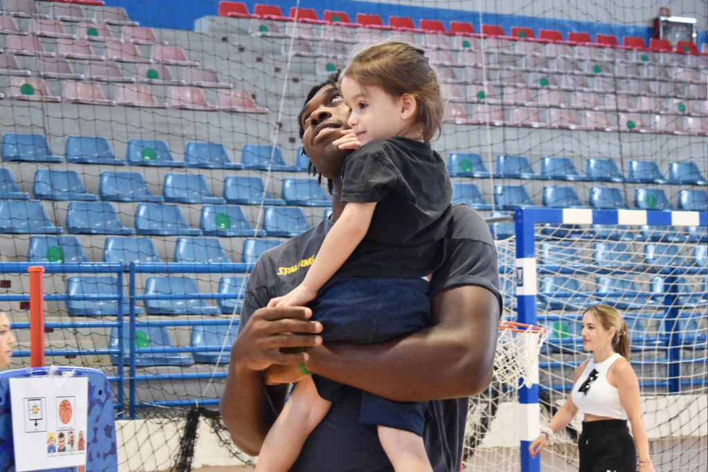 Entrenador sostiene a una niña mientras ambos sonríen en el polideportivo.