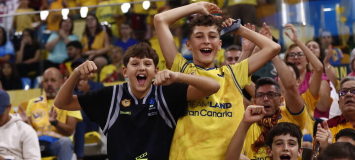 Niños animando en un partido, con público de fondo vestido de amarillo.